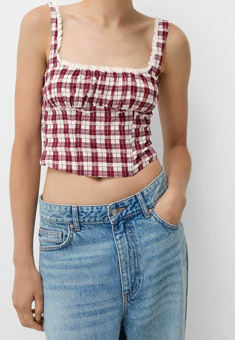 Femme portant un crop top sans manches à carreaux rouges et blancs avec bordure en dentelle, associé à un jean bleu taille haute, une main dans la poche.