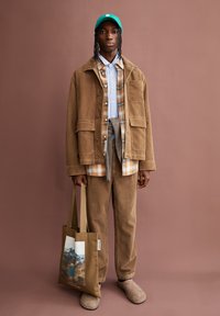 Brown corduroy jacket, plaid button-up shirt over a light blue top, matching brown corduroy pants, and beige slippers. Green cap and tote bag featured.