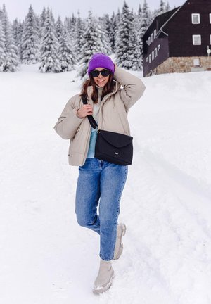 Béžová péřová bunda, světle modrý svetr, modré džíny a šedé boty. Černá kabelka přes rameno s klopou. Fialová pletená čepice a sluneční brýle. Sněhové pozadí.