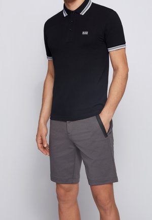 Mann trägt ein schwarzes Hugo Boss Polo mit weiß gestreiftem Kragen und Ärmeln, kombiniert mit grauen Shorts mit schwarzem Besatz an den Taschen.