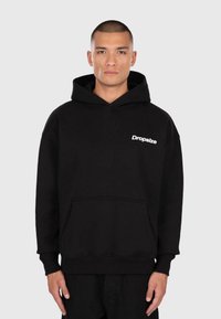 Jonge man met kort haar, gekleed in een zwarte hoodie met witte "Dropsize" tekst, staand tegen een effen lichtgrijze achtergrond.
