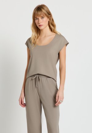 Giovane donna con capelli biondi che indossa una maglietta taupe a maniche corte e pantaloni coordinati con coulisse, su sfondo semplice.