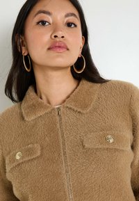 Veste zippée marron et duveteuse avec un col, ornée de boutons dorés sur les poches poitrine et d'une texture douce.