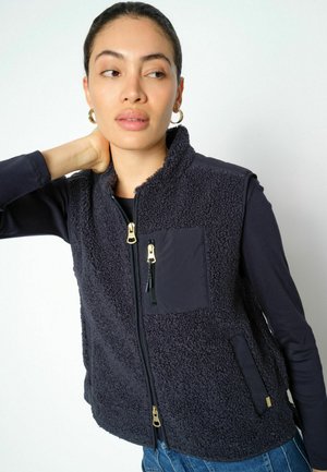 Woman wearing dark fleece zip-up vest over long sleeve shirt, looking sideways with hand behind neck, on plain light background.