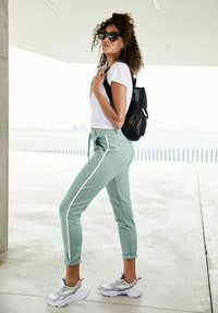 Mintgrüne Jogginghosen mit weißen Seitenstreifen, ein weißes, eng anliegendes T-Shirt und klobige weiße Sneaker. Inklusive eines schwarzen Mesh-Rucksacks.