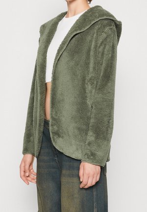 Persona que lleva una chaqueta con capucha abierta y peluda de color verde oliva sobre un top corto blanco y vaqueros anchos oscuros, de pie frente a un fondo sencillo.
