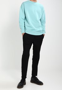 Sudadera de color teal claro con cuello redondo y puños acanalados, combinada con pantalones negros ajustados y zapatos negros con cordones, de pie sobre un fondo neutro.