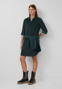Abito camicia in cotone verde scuro con bottoni sul davanti, maniche lunghe e tasche laterali. Stylisticamente abbinato a un top a righe blu navy e un maglione verde annodato in vita.