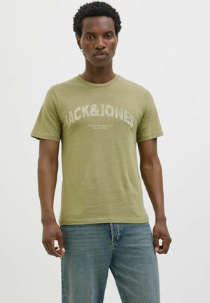 T-shirt en coton vert olive avec un grand logo blanc "JACK&JONES" et un texte plus petit, à manches courtes et col rond. Porté avec un jean bleu.