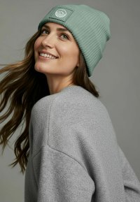 Green ribbed beanie with a sewn-on circular logo, worn with a light gray textured sweater. Hair falls softly over one shoulder.