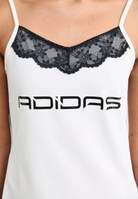 Bela majica na naramnice s črno čipko in valovitim robom, z velikim črnim logotipom "ADIDAS" natisnjenim spredaj v moderni pisavi.