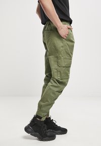 Pantalones cargo de color verde olivo con cintura elástica, bolsillos laterales y puños elásticos. Llevados con calzado negro con suela gruesa.