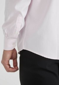 Chemise rose clair à manches longues avec un poignet boutonné et un ourlet droit, associée à un pantalon noir. Texture lisse, design à coupe classique.