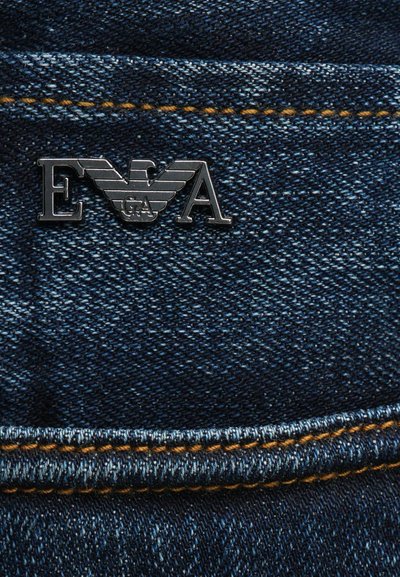 Σκούρο μπλε τζιν με υφή στην επιφάνεια, που διαθέτει αντίθετα πορτοκαλί ραφές και ένα ασημένιο λογότυπο E.A. με φτερά μπροστά.