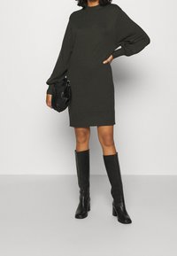 Vestito in maglia verde scuro con collo alto e maniche a sbuffo, abbinato a stivali neri con tacco fino al ginocchio. Accessori comprendono una borsa nera.