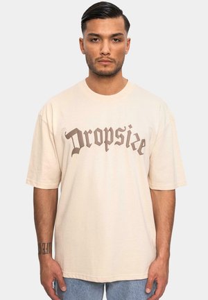 Beige bomull t-skjorte med "Dropsie" trykt i brun gotisk skrift. Avslappet passform med korte ermer og rund hals.