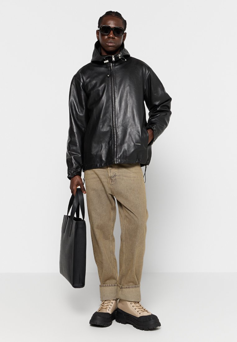 Zwarte leren jas met een capuchon, rits sluiting en verstelbare zoom, gecombineerd met beige broek en zwarte laarzen, met een zwarte tote bag.