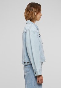 Chaqueta de mezclilla azul claro con un corte ligeramente cropped, frente de botones, bolsillos en el pecho y herrajes de metal. Textura suave y diseño casual.