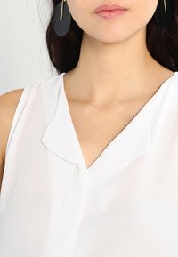 Witte, mouwloze blouse met een zachte, vloeiende textuur en een subtiele kraag. Grote zwarte ronde oorbellen hangen aan de oorlellen.