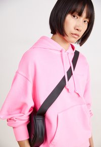 Felpa rosa con cappuccio regolabile, tasca frontale e polsini a coste. Indossata con una borsa a tracolla nera con texture liscia e tracolla.