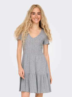 Lachende vrouw met lang krullend blond haar, gekleed in een grijze geribbelde gebreide jurk met korte mouwen en een gelaagde rok.