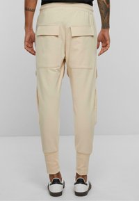 Beige cargo bukser med to øverste baglommer, elastisk talje og ribbede manchetter, parret med sort og hvide sko.