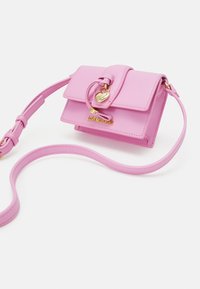 Růžová kožená kabelka se strukturovaným designem, horním zavíráním na klopě, zlatými kovovými doplňky a odnímatelným popruhem. Obsahuje přívěsek a logo značky.