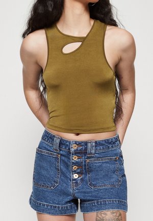 Frau trägt ärmelloses olivgrünes Crop-Top mit Ausschnitt und hoch taillierte blaue Jeansshorts mit vier Knöpfen und sichtbarem Tattoo am Oberschenkel.