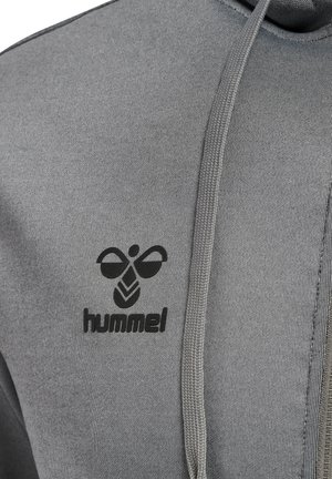 Nahaufnahme von grauem Hoodie-Stoff mit schwarzem hummel-Bienenlogo und Markennamen nahe der Brust, mit Kordelzug daneben hängend.