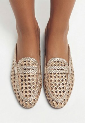 Pieds portant des mocassins tissés en or métallique avec garniture tressée sur un fond blanc uni, vus de dessus.