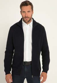 Marinblå zip-up kofta i ribbad stickning, med hög krage, långa ärmar och sidofickor med dragkedja. Bärs över en vit skjorta.