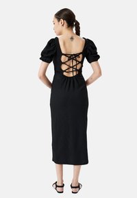 Vestido midi negro con mangas abullonadas, tejido texturizado, espalda descubierta con detalle de lazada y dobladillo sencillo. Llevado con sandalias negras de tiras.