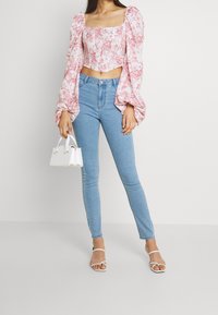 Top court blanc avec motif floral rose, manches bouffantes, associé à un jean skinny bleu clair taille haute. Accessoirisé avec un petit sac à main blanc.