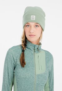 Berretto in maglia verde chiaro con risvolto e patch del logo; abbinato a una giacca verde con zip che presenta un tessuto strutturato e tasca laterale.