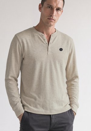 Mand iført beige langærmet henley-shirt med tre knapper, hænderne i lommerne, stående mod en ensfarvet lys baggrund.