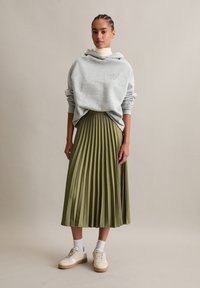 Frau trägt einen grauen oversized Hoodie, einen olivgrünen, plissierten Midirock, weiße Socken und beigen Sneakers und steht vor einem schlichten Hintergrund.