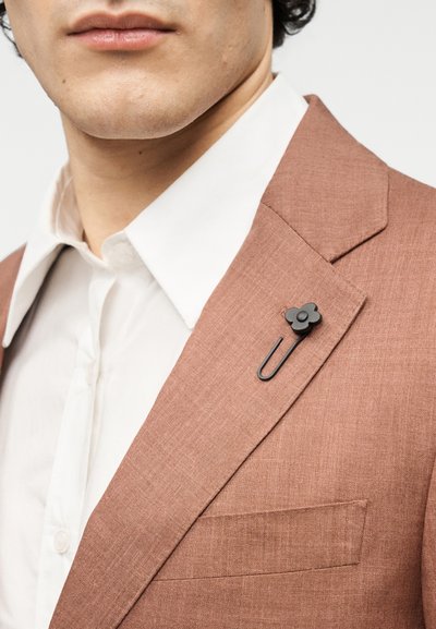 Ανοιχτό καφέ σακάκι με υφή και εγκοπτή πέτο; διαθέτει μαύρο λουλουδάτο καρφίτσα πέτου και λευκό πουκάμισο με γιακά από κάτω.