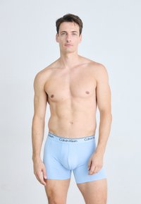 Kaalgebekte man met kort bruin haar, gekleed in lichtblauwe Calvin Klein boxer shorts, staande tegen een effen lichte achtergrond.