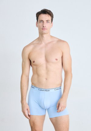 Uomo a torso nudo con capelli castani corti che indossa boxer blu chiaro di Calvin Klein, in piedi contro uno sfondo semplice e chiaro.