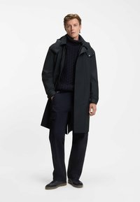 Impermeabile trench coat nero con colletto alto, cerniera frontale e chiusure a scatto. Indossato sopra un maglione navy a trama e pantaloni cargo.
