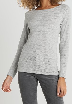 Femme portant un haut en maille texturée à manches longues gris clair et un pantalon gris foncé, une main détendue le long du corps.