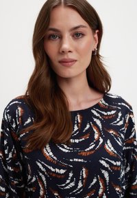 Młoda kobieta z długimi brązowymi włosami, nosząca srebrne kolczyki w kształcie kół oraz granatową bluzkę z białymi i rdzawymi abstrakcyjnymi wzorami, z neutralnym wyrazem twarzy, na białym tle.