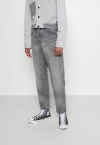 Jeans de denim gris con corte recto, combinados con una camisa de button-up gris claro que presenta un bolsillo oscuro, y zapatillas negras de caña alta.