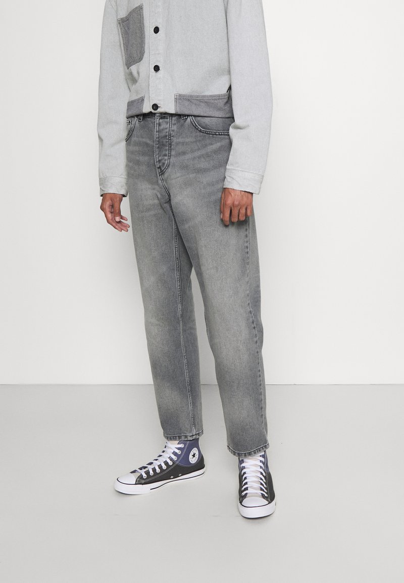 Jeans de denim gris con corte recto, combinados con una camisa de button-up gris claro que presenta un bolsillo oscuro, y zapatillas negras de caña alta.