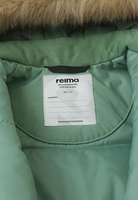 Interno di una giacca imbottita verde menta con cappuccio peloso, presenta un'etichetta di cura con la taglia 92/2T e un messaggio che incoraggia a passare l'articolo ad altri.
