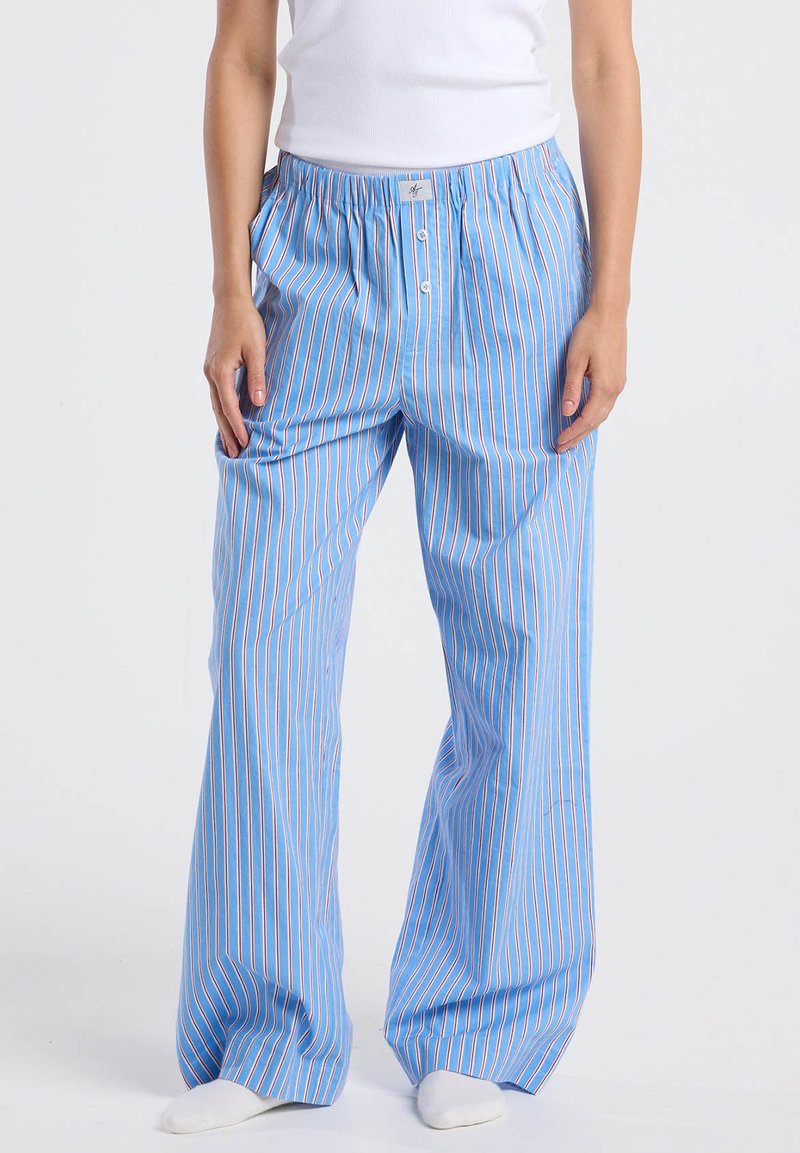 Henkilö, jolla on yllään löysät, siniset pyjamahousut, joissa on pystysuorat punaiset ja valkoiset raidat, valkoiset sukat ja valkoinen hihaton toppi.