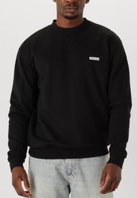 Svart sweatshirt med rund hals, ribbade muddar och nederkant. Har en vit "WOODBIRD" logotyp på bröstet, tillverkad av ett slätt tyg.