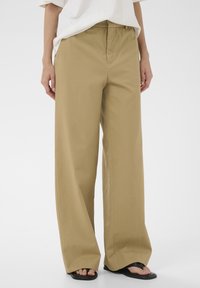 Pantalon beige à jambes larges porté avec un haut blanc et des sandales en caoutchouc noires, visible de la taille aux pieds sur un fond blanc.