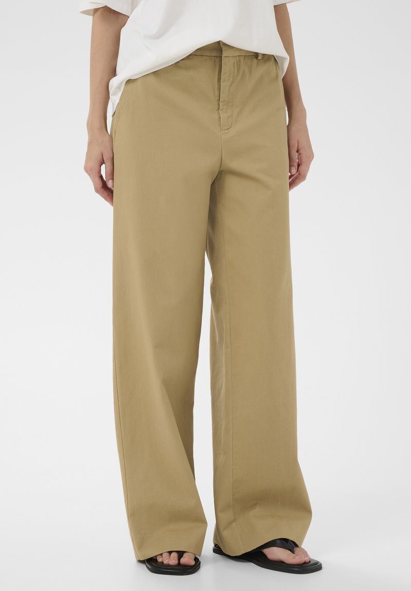Pantalon beige à jambes larges porté avec un haut blanc et des sandales en caoutchouc noires, visible de la taille aux pieds sur un fond blanc.