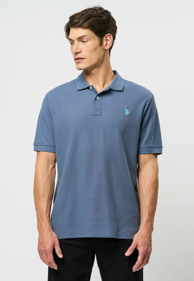Polo tričko v tlumené modré tkanině, s klasickým límcem, krátkými rukávy, dvouknoflíkovou légou a malým vyšitým logem na hrudi.